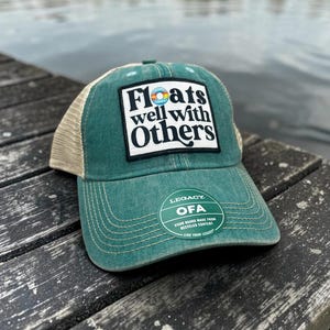 May include: A green baseball cap with a tan mesh back and a curved brim. The cap features the text "Floats well with Others" on a rectangular patch. A circular sticker on the brim reads "Legacy OFA" and "Visor board made from recycled content."