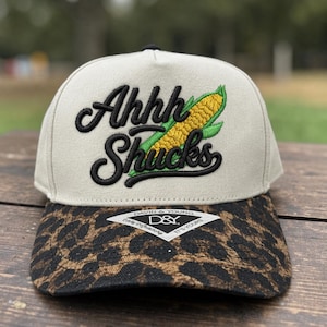 May include: A beige and black baseball cap with the text "Ahhh Shucks" embroidered in black script, along with a corn graphic. The brim features a leopard print design. The cap has a black top button and a tag that says "David & Young".