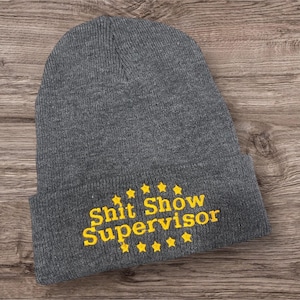 May include: A gray beanie with the embroidered text "Shit Show Supervisor" in yellow, along with yellow star accents. The hat is made of a knitted material and is displayed on a wooden surface.