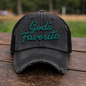 May include: A distressed charcoal gray baseball cap with a black mesh back and frayed edges on the brim. The front panel features the text God's Favorite embroidered in a raised, dark green script font with a distinct three-dimensional appearance.
