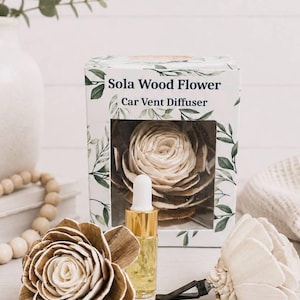 May include: A Sola Wood Flower Car Vent Diffuser set featuring a white box with green leaf illustrations, a small glass bottle with a gold cap and white dropper, and two cream-colored wooden flowers, one with a black plastic car vent clip attached.