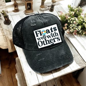 May include: A distressed black trucker hat with a mesh back and a curved brim. The hat features a white square patch with the text "Floats well with Others" and a colorful graphic of a life preserver.