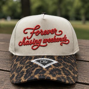 May include: A beige baseball cap featuring the red embroidered text Forever chasing weekends on the front panel. The brim displays a brown and black leopard print pattern and includes a white diamond-shaped sticker with the text David & Young D&Y.