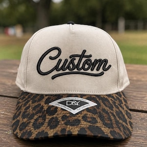 May include: A beige baseball cap featuring the word Custom embroidered in black script on the front panel. The brim displays a brown and black leopard print pattern and includes a white diamond-shaped sticker with the text David & Young D&Y.