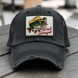 May include: A charcoal gray distressed trucker hat featuring a rectangular patch with an embroidered green fish wearing a yellow cowboy hat and glasses, accompanied by the red cursive text "Well Sheeyit" on a white background.