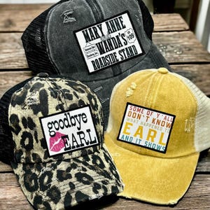 May include: Three distressed trucker hats with mesh backs. One is leopard print with "goodbye EARL" text. Another is gray with "MARY ANNE WANDA'S ROADSIDE STAND" text. The third is yellow with "SOME OF Y'ALL DON'T KNOW WHAT HAPPENED TO EARL AND IT SHOWS" text.