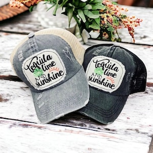 May include: Two distressed baseball caps with the text "Tequila, lime, and sunshine" on a white patch. One cap is gray with a tan mesh back, the other is dark gray with a black mesh back. The caps are on a white wooden surface.
