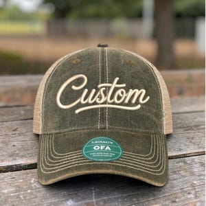 May include: An olive green baseball cap with a mesh back and a distressed finish. The front panel features the word Custom embroidered in a cream-colored script font. A green circular sticker on the brim displays the text Legacy OFA.