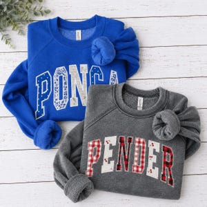May include: A royal blue sweatshirt with the word PONCA in white and blue floral applique letters and a charcoal gray sweatshirt with the word PENDER in white, red, and black plaid applique letters featuring small cherry illustrations.