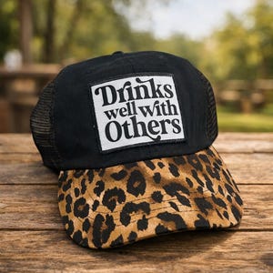 May include: Black trucker hat with a leopard print brim. The hat features a white square patch with the words "Drinks well with Others" in a stylized font. The hat is sitting on a wooden surface.