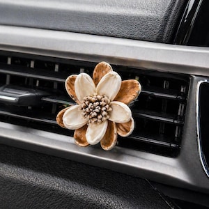 May include: A flower-shaped car air freshener with cream and brown petals and a textured center. The air freshener is attached to a car vent, with a black and silver dashboard visible in the background.