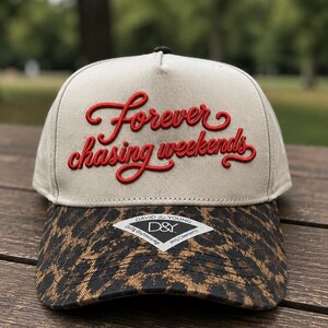 May include: A beige baseball cap featuring red embroidered cursive text that reads Forever chasing weekends. The brim displays a brown and black leopard print pattern and includes a white diamond-shaped sticker with the text David & Young D&Y.