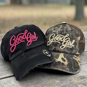 May include: Two baseball caps with mesh backs. The black cap features distressed detailing and the text Good Girl embroidered in bright pink. The second cap has a brown and black leopard print pattern with the text Good Girl in cream embroidery.