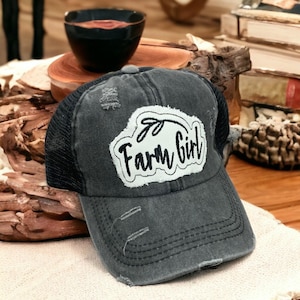 May include: A charcoal gray distressed baseball cap with a black mesh back. The front features a white fabric patch with frayed edges, displaying the black embroidered text Farm Girl and a small leaf graphic above the lettering.