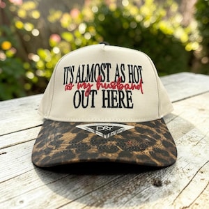 May include: A beige baseball cap with a brown and black leopard print brim. The front panel features black embroidered text that reads IT'S ALMOST AS HOT OUT HERE with the red embroidered text as my husband in the center. A D&Y sticker is on the brim.