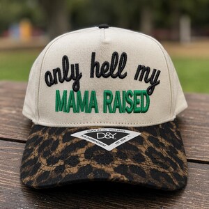 May include: A beige baseball cap with a leopard print brim. The front features black embroidered text that reads only hell my and green embroidered text that reads MAMA RAISED. A diamond-shaped David & Young brand sticker is attached to the brim.