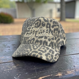 May include: A baseball cap featuring a grey and black leopard print pattern. The front of the cap displays the embroidered text "forever chasing weekends" in a light cream-colored cursive script font across the center of the crown.
