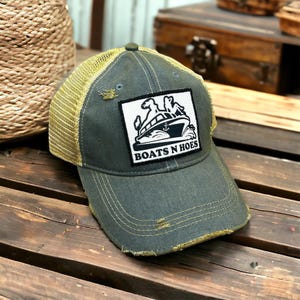 May include: A distressed olive green trucker hat with a khaki mesh back. The front features a white square patch with a black and white boat graphic and the text "BOATS N HOES". The hat has a curved brim and a button on top.