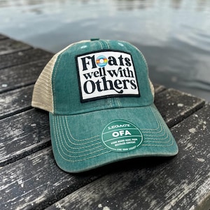 May include: A green and beige trucker hat featuring a white rectangular patch with the black text Floats well with Others and a small colorful circular icon. A round sticker on the brim displays the text Legacy OFA Visor board made from recycled content.