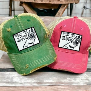 May include: Two distressed trucker hats, one green and one pink, featuring a white rectangular patch with a black line drawing of a chicken and the text "WHAT THE CLUCK". Both hats have mesh back panels and frayed edges on the front bill.
