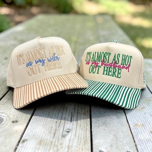 May include: Two beige baseball caps with embroidered text. The left cap features tan and white striped brim with text IT'S ALMOST AS HOT as my wife OUT HERE. The right cap has green and white striped brim with text IT'S ALMOST AS HOT as my husband OUT HERE.