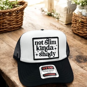May include: A black and white trucker hat with the text "not slim kinda shady" on the front. The hat has a black brim and mesh back. The hat also has a tag that says "OTTO World Famous Foam Front OSFM Trucker Durable Snap Closure".