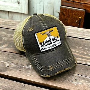 May include: A distressed charcoal gray baseball cap with a tan mesh back and a rectangular patch on the front. The patch features a yellow background, a black bull skull illustration, and the text RAISIN HELL WITH THE HIPPIES AND THE COWBOYS.