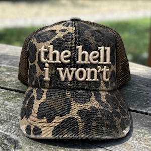 May include: A brown and black leopard print trucker hat featuring a mesh back and a curved brim. The front panel displays the embroidered text "the hell i won't" in a light tan, sans-serif font with a raised, textured appearance.