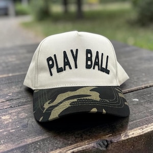 May include: A beige baseball cap with the words "PLAY BALL" in bold black letters across the front. The brim features a camouflage pattern in shades of green, brown, and black. The cap is sitting on a wooden surface.
