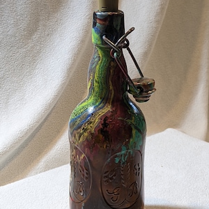 May include: A glass bottle with a colorful abstract paint design. The bottle has a metal swing top lid and a cork stopper. The bottle has a brown label with a design of wheat stalks and the letter "B".