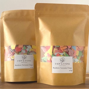 May include: Two brown paper bags with a clear window displaying colorful terrazzo chips. Each bag has a white label with the text "STEM & STONE DESIGNS Rainbow Terrazzo Chips 200g". The chips are various colors.