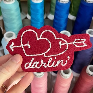 Op de afbeelding: Een rode vilt patch met twee harten doorboord door een pijl, geborduurd met witte draad. Het woord "darlin'" is eronder gestikt. De achtergrond toont spoelen met kleurrijk garen.
