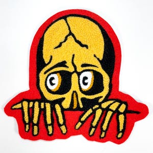 May include: An embroidered patch featuring a yellow skull with black eye sockets and white pupils, positioned above two yellow skeletal hands with black outlines. The entire design is set against a vibrant red background border.