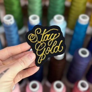 May include: A black embroidered patch with the words "Stay Gold" in gold thread. The patch has a unique cloud-like shape. The background features spools of colorful thread, suggesting a crafting or sewing theme.