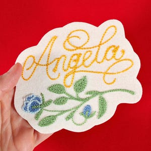 K&ouml;nnte beinhalten: Gestickter Aufn&auml;her mit dem Namen "Angela" in goldener Schrift, verziert mit einem gr&uuml;nen und blauen Blumenmuster. Der Aufn&auml;her wird vor einem roten Hintergrund pr&auml;sentiert, der die detaillierten Stickereien und den Farbkontrast hervorhebt.