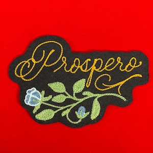 K&ouml;nnte beinhalten: Schwarzer, gestickter Aufn&auml;her mit dem Wort "Prospero" in goldener Schrift. Der Aufn&auml;her zeigt ein Blumendesign mit gr&uuml;nen Bl&auml;ttern und einer blauen Blume. Der Aufn&auml;her ist vor einem leuchtend roten Hintergrund platziert.