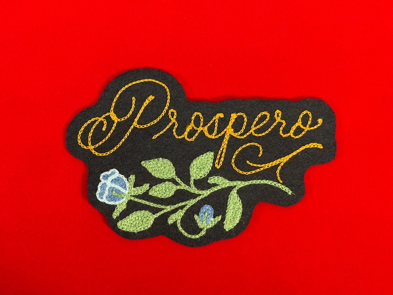 K&ouml;nnte beinhalten: Schwarzer, gestickter Aufn&auml;her mit dem Wort "Prospero" in goldener Schrift. Der Aufn&auml;her zeigt ein Blumendesign mit gr&uuml;nen Bl&auml;ttern und einer blauen Blume. Der Aufn&auml;her ist vor einem leuchtend roten Hintergrund platziert.