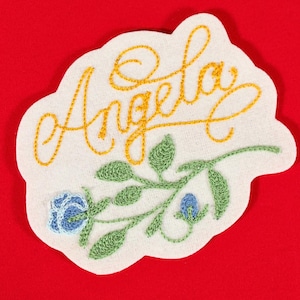 Puede incluir: Parche bordado con el nombre "Angela" en escritura dorada. El parche presenta una flor azul y hojas verdes sobre un fondo blanco, todo cosido con hilo. El parche está sobre un fondo rojo.