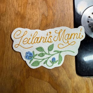 K&ouml;nnte beinhalten: Gestickter Aufn&auml;her mit dem Text "Leilani's Mami" in goldfarbenem Faden. Der Aufn&auml;her zeigt ein Blumendesign mit gr&uuml;nen Bl&auml;ttern und blauen Bl&uuml;ten auf wei&szlig;em Grund. Der Aufn&auml;her liegt auf einer Holzoberfl&auml;che.