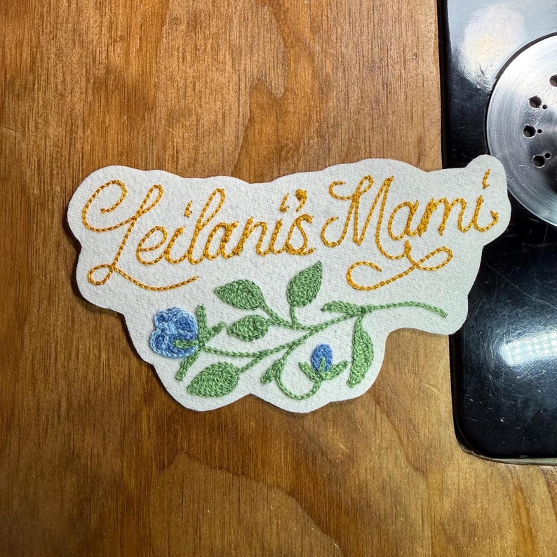 K&ouml;nnte beinhalten: Gestickter Aufn&auml;her mit dem Text "Leilani's Mami" in goldfarbenem Faden. Der Aufn&auml;her zeigt ein Blumendesign mit gr&uuml;nen Bl&auml;ttern und blauen Bl&uuml;ten auf wei&szlig;em Grund. Der Aufn&auml;her liegt auf einer Holzoberfl&auml;che.