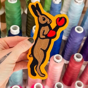 Può includere: Una toppa ricamata raffigurante un coniglio marrone in piedi sulle zampe posteriori, che indossa guanti da boxe rosso vivo. Il coniglio ha contorni neri ed è posizionato su uno sfondo in feltro giallo brillante, creando un design grafico audace.