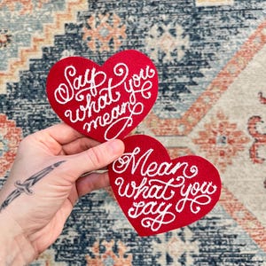 hand holding 2 red heart patches that say "Say what you mean" and "Mean what you say" in fancy cursive script
