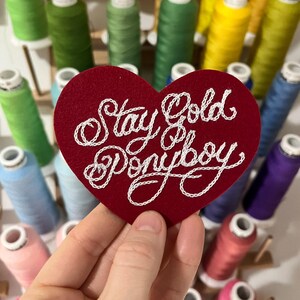 hand holding red custom felt heart patch that says "Stay Gold Ponyboy" in fancy cursive script