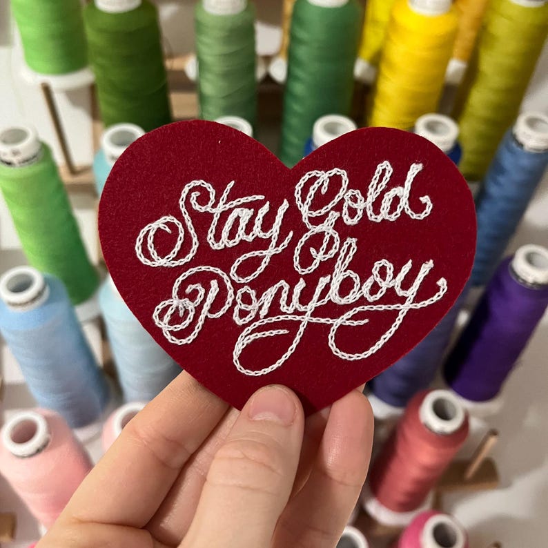 hand holding red custom felt heart patch that says "Stay Gold Ponyboy" in fancy cursive script