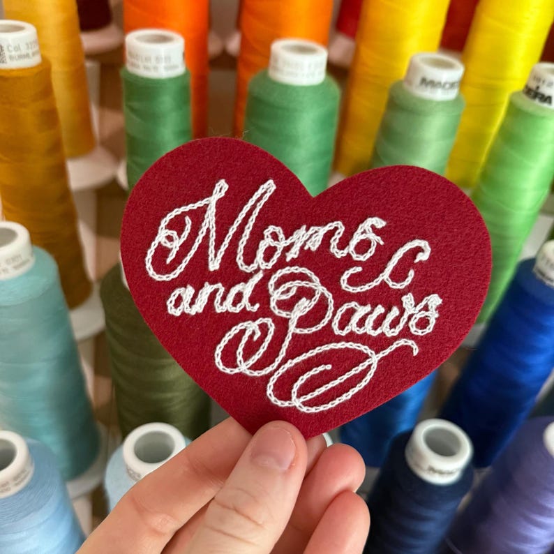 May include: A heart-shaped red felt patch featuring white embroidered cursive text that reads Moms and Paws. The patch is held against a background of various colorful spools of sewing thread in shades of yellow, orange, green, blue, and purple.