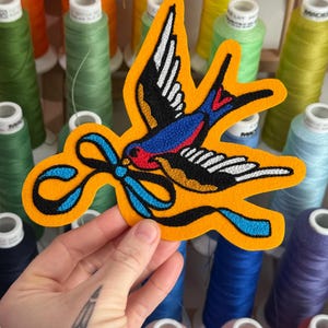 May include: An embroidered patch featuring a swallow bird in flight, rendered in black, blue, red, and white thread. The bird holds a bright blue ribbon in its beak, all set against a vibrant orange felt background with a die-cut border.