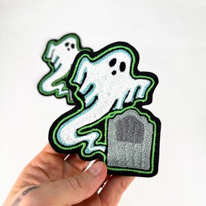 May include: Embroidered patch featuring a white ghost with a green outline and a gray tombstone. The patch has a black border. The design is on a white background. The patch is held in a hand.