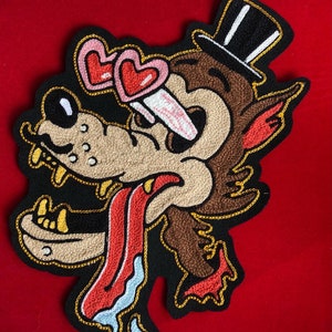 Könnte beinhalten: Gestickter Aufnäher mit einem Cartoon-Wolf mit herzförmiger Sonnenbrille und Zylinder. Der Wolf hat ein braunes und beiges Gesicht mit roter Zunge und blauem Sabber. Der Aufnäher ist mit goldenem Faden auf schwarzem Hintergrund umrandet.