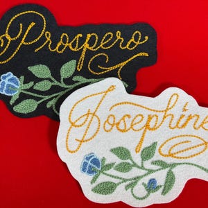 K&ouml;nnte beinhalten: Zwei gestickte Namensaufn&auml;her. Einer ist schwarz mit dem Namen "Prospero" in goldener Schrift, der andere ist wei&szlig; mit "Josephine" in goldener Schrift. Beide haben florale Akzente mit blauen Blumen und gr&uuml;nen Bl&auml;ttern.