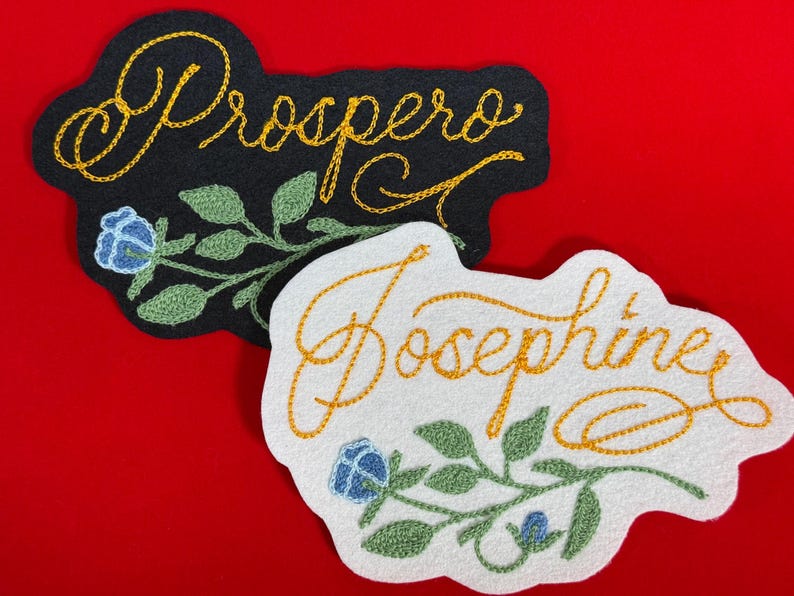 K&ouml;nnte beinhalten: Zwei gestickte Namensaufn&auml;her. Einer ist schwarz mit dem Namen "Prospero" in goldener Schrift, der andere ist wei&szlig; mit "Josephine" in goldener Schrift. Beide haben florale Akzente mit blauen Blumen und gr&uuml;nen Bl&auml;ttern.